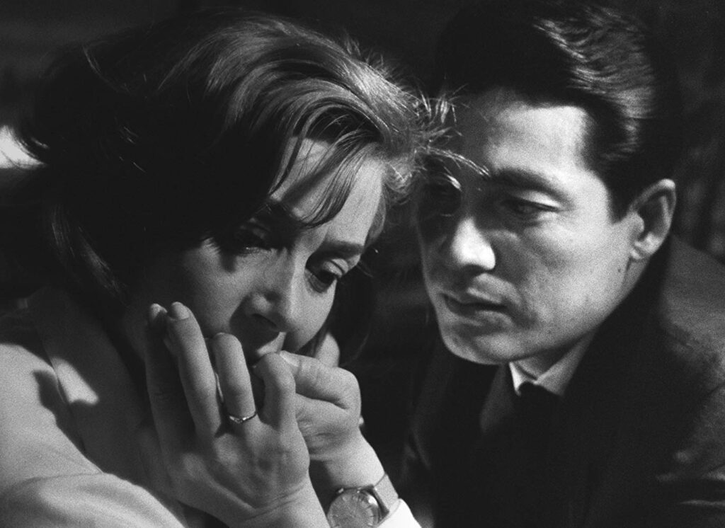 "Filmstill uit Alain Resnais' Hiroshima Mon Amour (1959): close-up van twee gezichten in intieme nabijheid tegen donkere achtergrond. De Franse vrouw en haar Japanse geliefde, gevangen in een moment van tedere omhelzing die tegelijkertijd de onoverbrugbare kloof belichaamt tussen wat zich laat zeggen en wat radicaal buiten symbolisering valt. Hun nabijheid contrasteert met de fundamentele onmogelijkheid van volledig begrip — een visuele metafoor voor Lacans conceptie van het reële als dat wat elk spreken overschrijdt, wat zich onttrekt aan elke poging tot representatie. In deze bevroren intimiteit kondigt zich af wat de psychoanalyse het trauma zal noemen: niet als gebeurtenis die zich laat verhalen, maar als terugkerende confrontatie met de grenzen van het zegbare zelf. De zwart-wit cinematografie versterkt het gevoel van een herinnering die zich tegelijkertijd opdringt en onttrekt — aanwezig als afwezigheid, zichtbaar als wat zich niet laat zien."
"Filmstill uit Hiroshima Mon Amour (1959): twee gezichten in intieme nabijheid, zwart-wit cinematografie. De Franse vrouw en haar Japanse geliefde belichamen de paradox van nabijheid en onmogelijkheid — een visuele metafoor voor wat zich onttrekt aan symbolisering." Michael Bijnens. Psychoanalyse Amsterdam.
